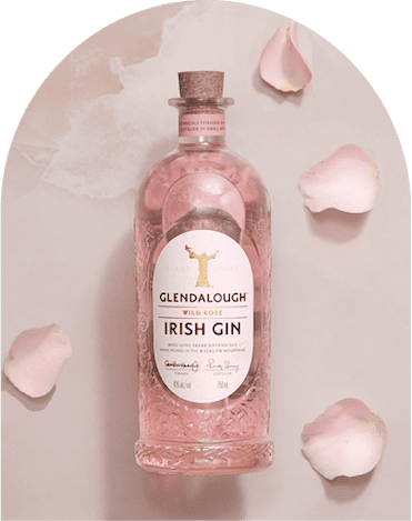 A bottle of Glendalough Wild Rose Irish Gin filled with pink liquid, surrounded by scattered pink rose petals against a soft pink arched background.