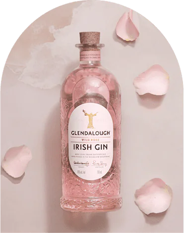 A bottle of Glendalough Wild Rose Irish Gin filled with pink liquid, surrounded by scattered pink rose petals against a soft pink arched background.