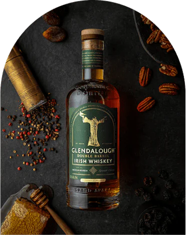A bottle of Glendalough Double Barrel Irish Whiskey on a dark stone surface, surrounded by honeycomb, peppercorns, pecans, and dried fruit.
